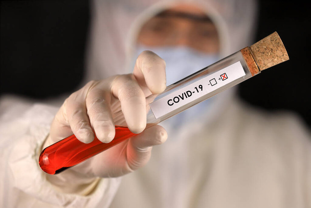 The doctor in a protective suit and mask holding a glass tube with a positive blood test result for the new rapidly spreading Coronavirus. Global coronavirus disease (COVID-19) 2020. - Foto, Imagen