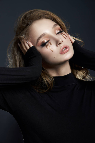 Beauty portrait blonde woman with makeup and big blue eyes, natural cosmetics, clean delicate skin of the girl face, black clothes. Drops of makeup trickle from the girl eyes - Photo, Image