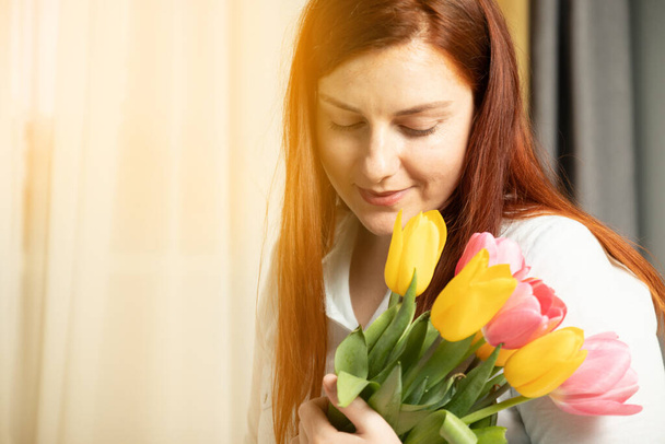 Frauentag am 8. März. Schöne Mädchen in einer blauen Baumwollbluse mit frischen Tulpen Strauß in ihren Händen, schnüffelt sie an einer grauen Wand Hintergrund - Foto, Bild