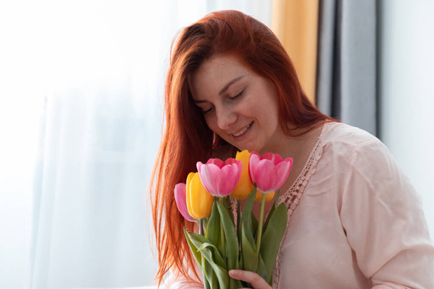 Hermosa jovencita de pelo largo sosteniendo un ramo de flores frescas de tulipanes en sus manos y olfateándolas en la habitación. Día de la Mujer 8 de marzo
 - Foto, Imagen