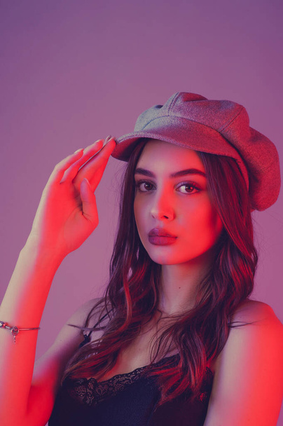 Glamorous young woman wears fashionable dress posing with fluttering hair. Girl in a black dress and a gray cap. girl with brown curly hairs. portrait. beautiful, stylish woman on a pink background - Φωτογραφία, εικόνα