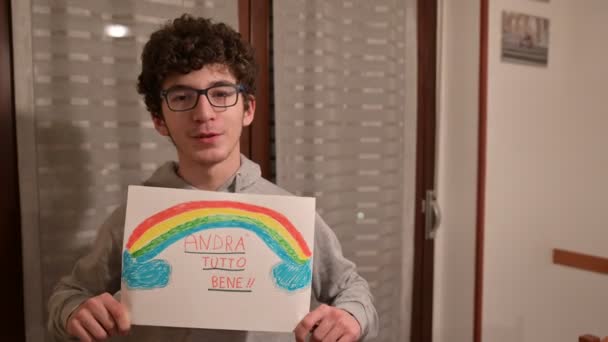 Turin, Piedmont, Italy. March 2020. Cornonavirus quarantine. Portrait of a caucasian boy with brown curly hair and glasses holding up a drawing with the rainbow and the message will be all right! - Footage, Video