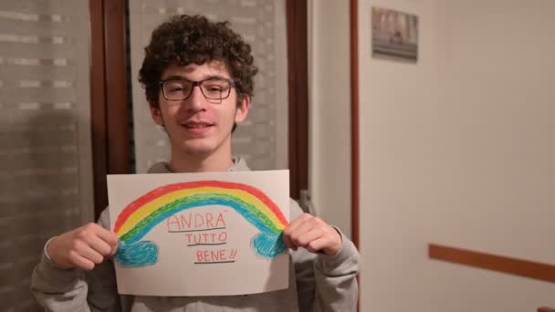 Turin, Piedmont, Italy. March 2020. Cornonavirus quarantine. Portrait of a caucasian boy with brown curly hair and glasses holding up a drawing with the rainbow and the message will be all right! - Footage, Video