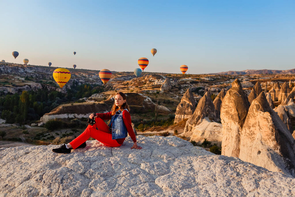 CAPPADOCIA, TURKEY  AUGUST 23, 2019: Young beautiful girl photographer with camera in her hands and flight of hot air balloons at sunrise in Cappadocia. Goreme, Cappadocia, Turkey - Foto, afbeelding