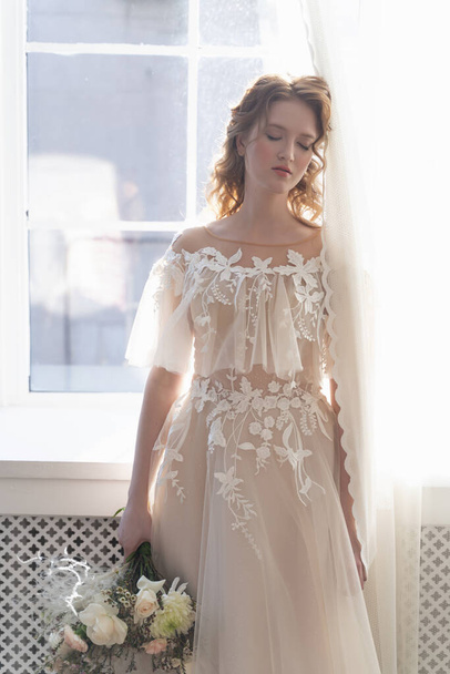 Красивая натуральная рыжая невеста, с голым макияжем, в белом платье, держит свадебный букет в руках, стоя у окна в светлом интерьере
 - Фото, изображение