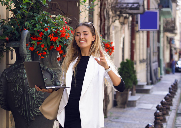 Amazing bright girl walking down the street with laptop in hand and speaking with someone via video chat. Spring warm day, blooming flowers outdoor. White smile, big lips, makeup and accessories. - Valokuva, kuva
