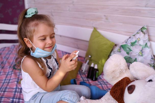 Blank grappig klein meisje met een medisch masker thuis doktertje spelen met een beer, een neusspray vasthouden. Quarantainebescherming tegen het coronavirus 2019-ncov. Isolatie thuis - Foto, afbeelding