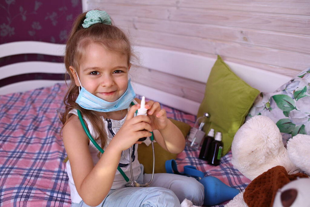 Blank grappig klein meisje met een medisch masker thuis doktertje spelen met een beer, een neusspray vasthouden. Quarantainebescherming tegen het coronavirus 2019-ncov. Isolatie thuis - Foto, afbeelding