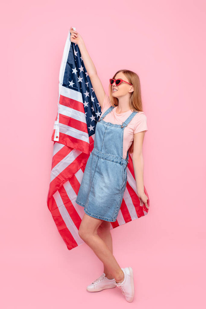 A young happy cheerful, stylish woman in sunglasses holds the US flag, standing on an isolated pink background - Valokuva, kuva