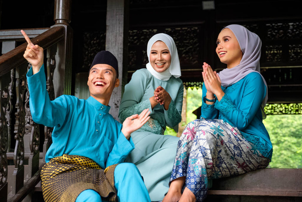 Grupa trzech muzułmanów malajskich w tradycyjnym stroju odbywają szczęśliwą rozmowę podczas uroczystości Aidilfitri na tarasie tradycyjnego drewnianego domu. Koncepcja mody Raya i muzułmańskiej. - Zdjęcie, obraz