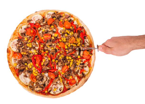Man 's hand met een pizzasnijder sneed de pizza geïsoleerd op witte achtergrond met clipping pad, bovenaanzicht. Italiaanse pizza met champignons, maïs, kerstomaten, courgettes en paprika 's of vegetarische vegetarische pizza - Foto, afbeelding