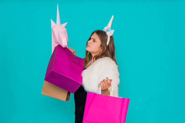 Portrait of a smiling pretty girl with shopping bags over blue background. Black friday concept. Kawaii teenage girl with unicorn horn buying a paper pink unicorn. - Photo, Image
