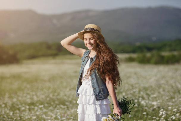Menina adolescente bonita em um vestido branco e chapéu andando em um campo de camomilas. Cabelo longo e saudável bonito. A ideia e conceito de saúde e alergias, cuidar de si mesmo
 - Foto, Imagem