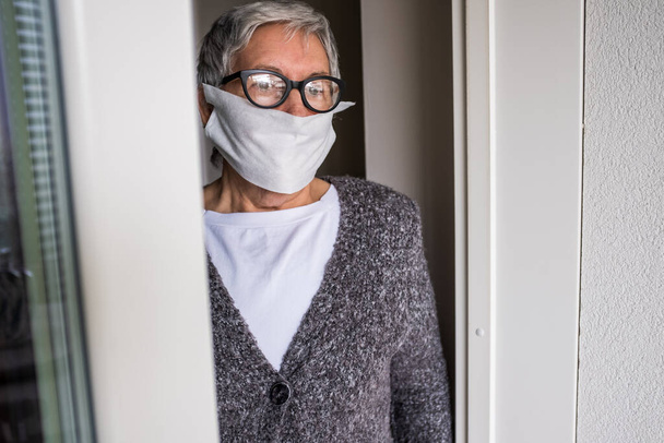 Besinnliche Seniorin steht am Fenster ihres Hauses und schaut hinaus. Vor allem ältere Menschen müssen geschützt werden, weil sie zu einer der Hauptrisikogruppen beim Ausbruch des Coronavirus gehören. - Foto, Bild