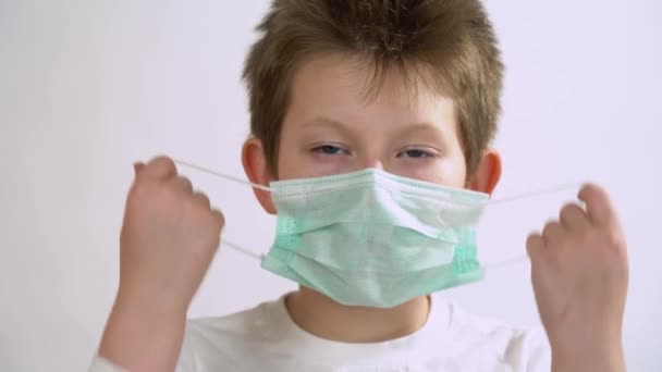 Ein kleines Kind als Arzt mit blauen Augen im langen Ärmel hebt ab und zieht sich eine Schutzmaske aus Nahsicht über das Gesicht. Kaukasisches Kind mit Verdacht auf 2019-nCoV-Infektion. Isolierter weißer Hintergrund. - Filmmaterial, Video