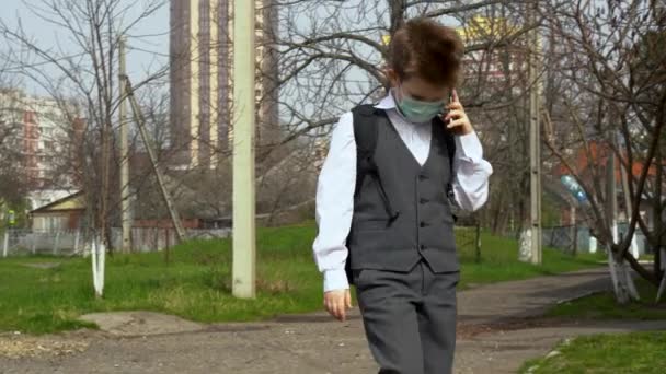Little boy with bag in suit, protective infection prevention mask walking, talking on phone against background of a school brick building. Child go to class despite the quarantine and isolation period - Footage, Video