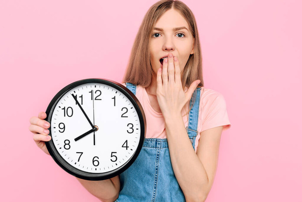 Shocked, a young lady on an isolated pink background holding a wall clock. - Φωτογραφία, εικόνα