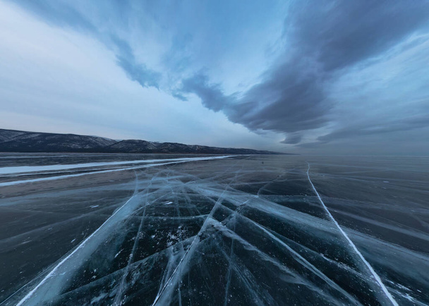Ice of Lake Baikal w pęknięciach w pobliżu wyspy Elenka o zachodzie słońca pod szarymi chmurami. Szeroka panorama - Zdjęcie, obraz