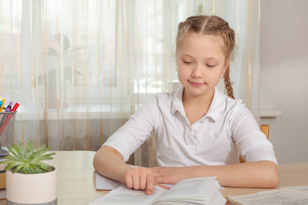 Schoolgirl studying at home. Young girl reading book at the table. Distance education, home education. Self isolation concept - Image - Fotoğraf, Görsel