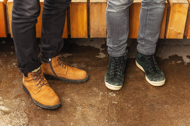 Braune und schwarze Winterschuhe an den Füßen. Füße in Schuhen aus nächster Nähe. Zwei Paar Füße in alten Stiefeln auf der Straße - Foto, Bild