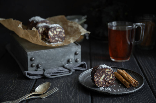 Stuk chocolade salami bestrooid met kokos- en kaneelstokjes op metalen plaat op donkere ondergrond - Foto, afbeelding