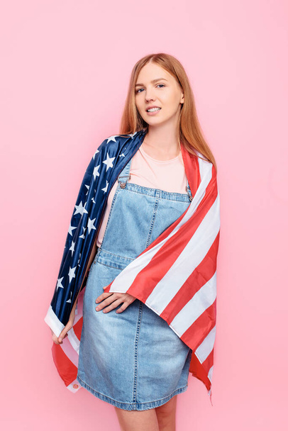 Photo of a young, fun, stylish woman holding a US flag while standing on an isolated pink background - Foto, afbeelding