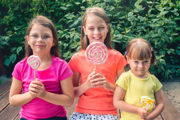 Tres niñas divertidas en camisetas coloridas con piruletas grandes - en la terraza del jardín
 - Foto, Imagen