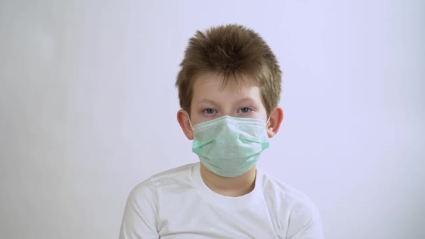 Caucasian pretty blonde kid with short hair in long sleeve wearing a green protective medical mask isolated on white background close up. The child has itchy nose and scratches it. Itching. - Footage, Video