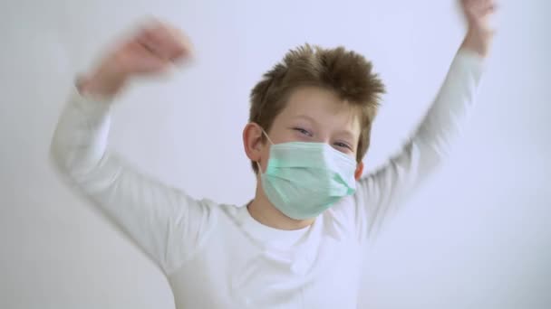 Pretty young blonde man in long sleeve wear protective medical mask on face from corona virus on white wall background close up. A happy little boy smile, dance, rejoices and looks at the camera. - Footage, Video
