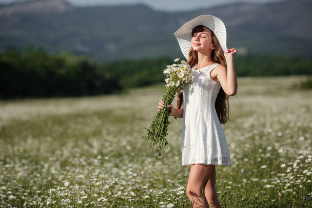 Beautiful cute teen girl on a walk in a daisy field, in a white dress and white hat. Idea and concept of happy growing up, health and allergy, skin and hair care. - Foto, Imagen