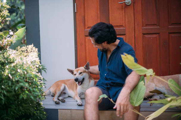 A man with dogs sits on the porch of a house in a tropical garden. A handsome brunette guy is resting on the steps among the tropical thickets near the house in the morning. - Foto, afbeelding
