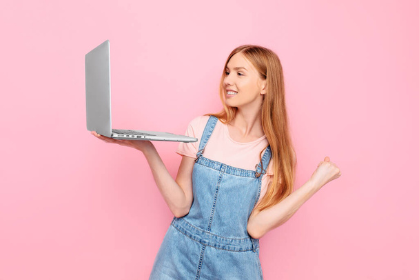 Portrait of a shocked young girl holding a laptop and showing a winning gesture isolated on a pink background - Foto, imagen