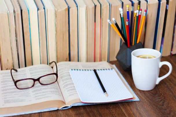 Open textbook, notebook, glasses, pencils in holder, cup of tea and stack of old book on wooden table, education concept background, many books piles with copy space for text - Photo, Image