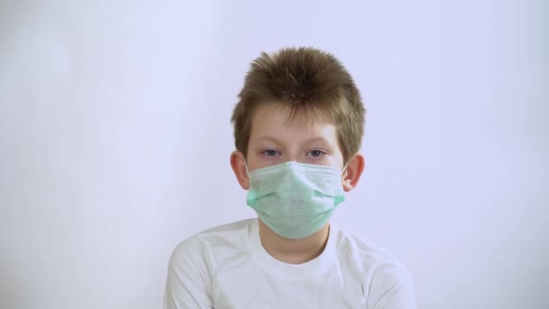 Attractive Caucasian boy with short blond hair in pullover in protective medical mask from corona virus nods his head up and down, say yes over a white wall background. Gesture of consent and agrees. - Footage, Video