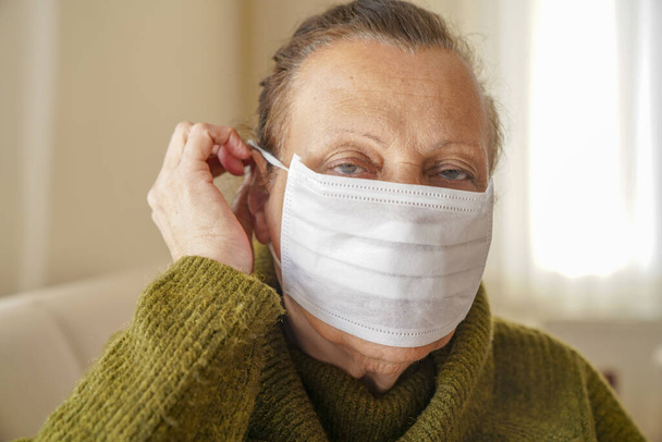 Портрет пожилой женщины в хирургической маске для защиты от коронного вируса. Бабушка сидит в своей гостиной и смотрит вверх
  - Фото, изображение