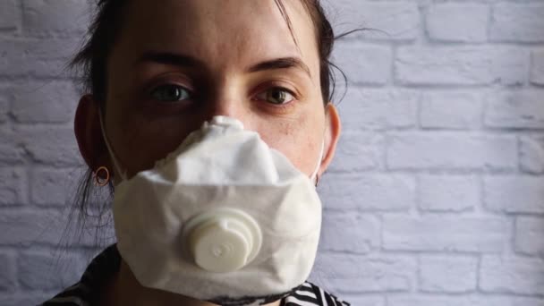 Portret van een jonge vrouw met een masker van een coronavirusepidemie tegen een witte bakstenen muur. Covid-19 gezondheids- en veiligheidsconcept, bescherming tegen het H1H1-virus. Begrepen, ruimte. opgewonden blik - Video