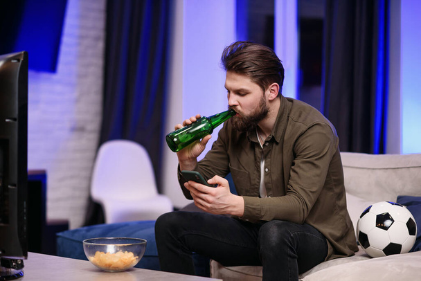 Slow motion of good-looking modern 30-aged guy with beard which sitting in front of tv and periodically watching football match and apps on his mobile while drinking beer - Foto, Imagen