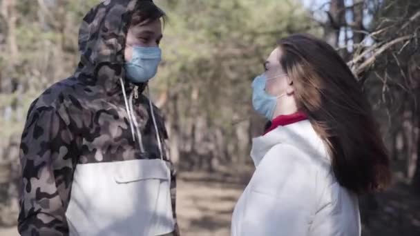 Side view of positive young Caucasian couple in face masks talking outdoors. Man and woman spending sunny spring day in park during coronavirus quarantine. Covid-19, lifestyle, pandemic, danger. - Πλάνα, βίντεο
