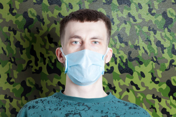 Ένας άνθρωπος με μια ιατρική μάσκα στο πρόσωπό του κατά τη διάρκεια μιας επιδημίας κορωναϊού, ενώ υπερασπίζεται τον εαυτό του ενάντια στη μόλυνση Covid-19 σε ένα πράσινο στρατιωτικό υπόβαθρο. - Φωτογραφία, εικόνα
