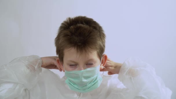 Beautiful young cute blonde kid with short hair in a protective suit put medical mask, respirator on his face, glasses, hood close up on white background. Quarantine by reason of coronavirus concept. - Imágenes, Vídeo