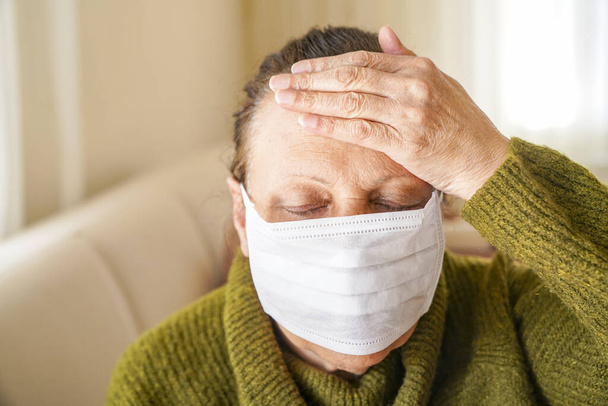 コロナウイルスに対する保護のために外科用マスクを身に着けている老婦人の肖像。祖母は彼女のリビングルームに座って検索します  - 写真・画像