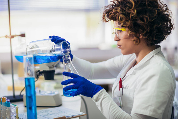 Joven científica atractiva en gafas protectoras y guantes usando tubo de ensayo con sonda de sustancia líquida azul en el laboratorio de investigación química científica
 - Foto, imagen