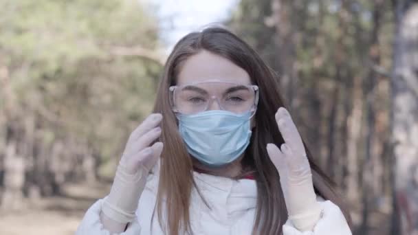 Young woman taking off protective eyeglasses and face mask and smiling at camera. Brunette Caucasian girl posing outdoors at the end of coronavirus quarantine. Covid-19 isolation, infectious disease. - Video