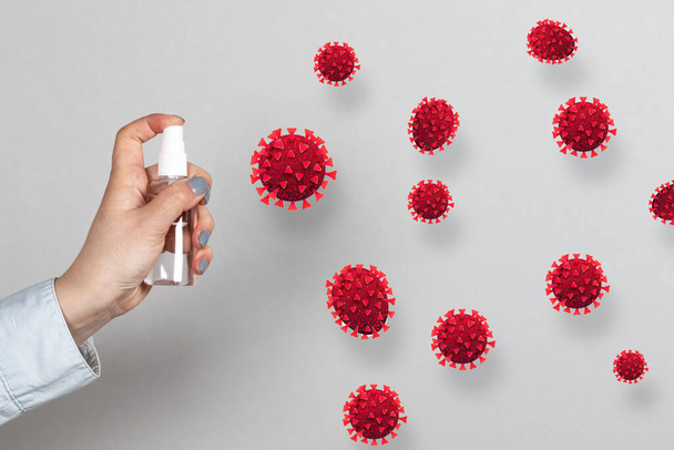 Weibliche Hand hält eine Flasche Antiseptikum und sprüht auf Coronavirus-Zellen auf grauem Wandhintergrund. - Foto, Bild