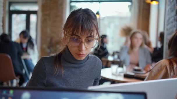 Νεαρή επιχειρηματίας με γυαλιά ηλίου που επικεντρώνεται στην οθόνη και δακτυλογράφηση σε φορητό υπολογιστή, ενώ κάθεται στο γραφείο στο χώρο εργασίας ή καφέ. - Πλάνα, βίντεο