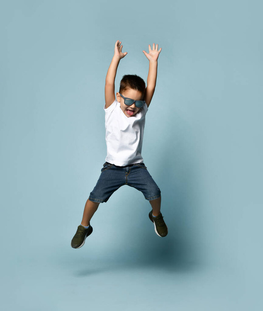 Little brunet child in sunglasses, white t-shirt, denim shorts and khaki sneakers. Jumping up with raised hands. Blue background - Fotó, kép
