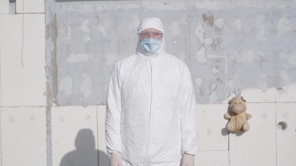 Caucasian boy in white safety suit, face mask and protective eyeglasses standing outdoors at the background of wall. Camera approaching to young Caucasian man walking out during Covid-19 quarantine. - Filmagem, Vídeo