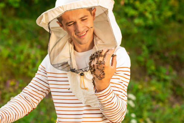 Happy beekeeper in a protective cap on the apiary. He looks at the hand, whole in bees. - Photo, Image