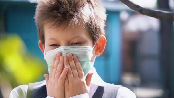 Child in protective medical mask hold his mouth with hands, cover face, look sad down. Nice blonde boy is afraid of getting infected. Kid recall sadly about good days and friends slow motion close up. - Footage, Video