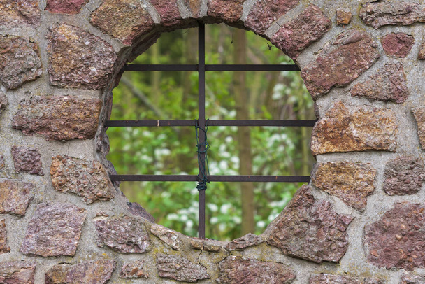 Yuvarlak deliğin pencere gibi, pencerenin de kafes gibi olduğu duvar. Parmaklıkların arkasında ormanın yeşil manzarası var. - Fotoğraf, Görsel